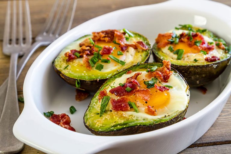 Baked avocado and eggs boats with bacon crumbles and chives sitting in white baking dish with two forks on the side