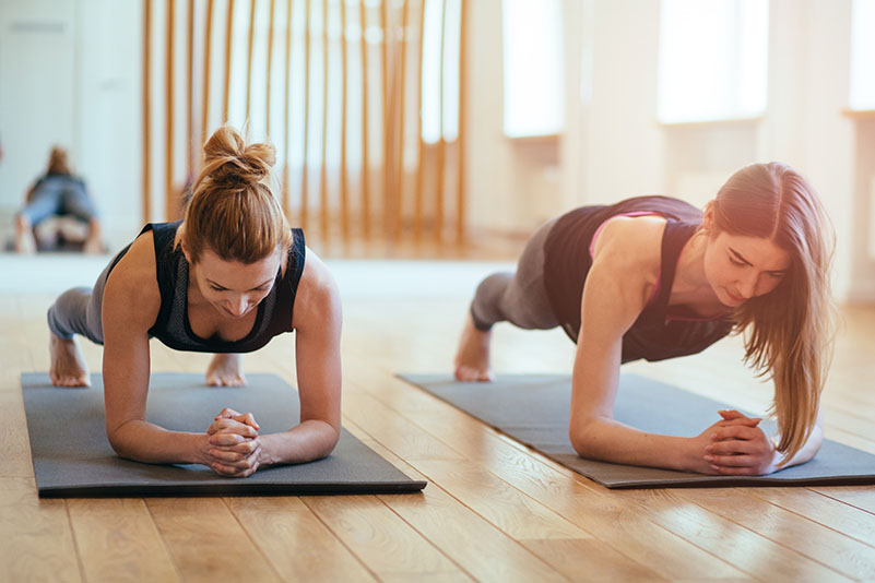 Front view of two sporty middle aged women doing planking exercise indoors together with a natural light in modern interior sport studio hall or yoga class.
